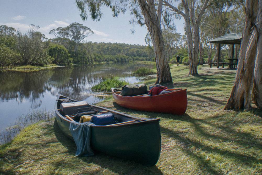 Canoes pulled up on a grassy bank beside dark still water fringed by paperbarks