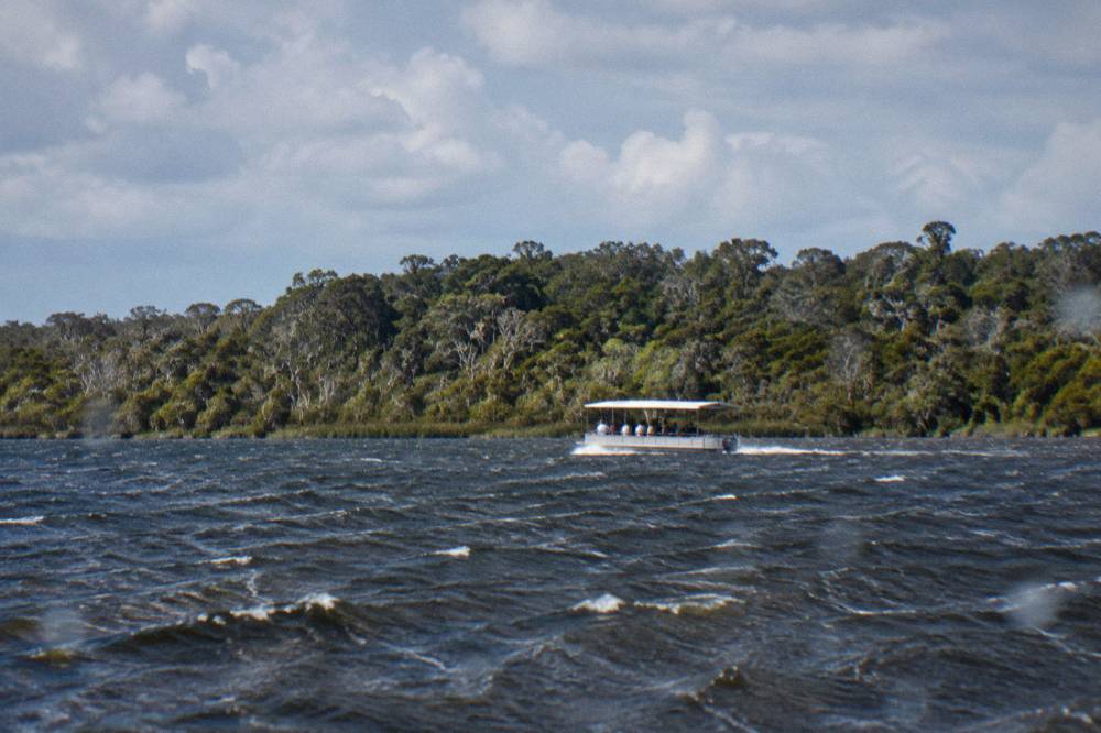 A small cruise boat travelling across a wide shallow lake toward distant forested shoreline