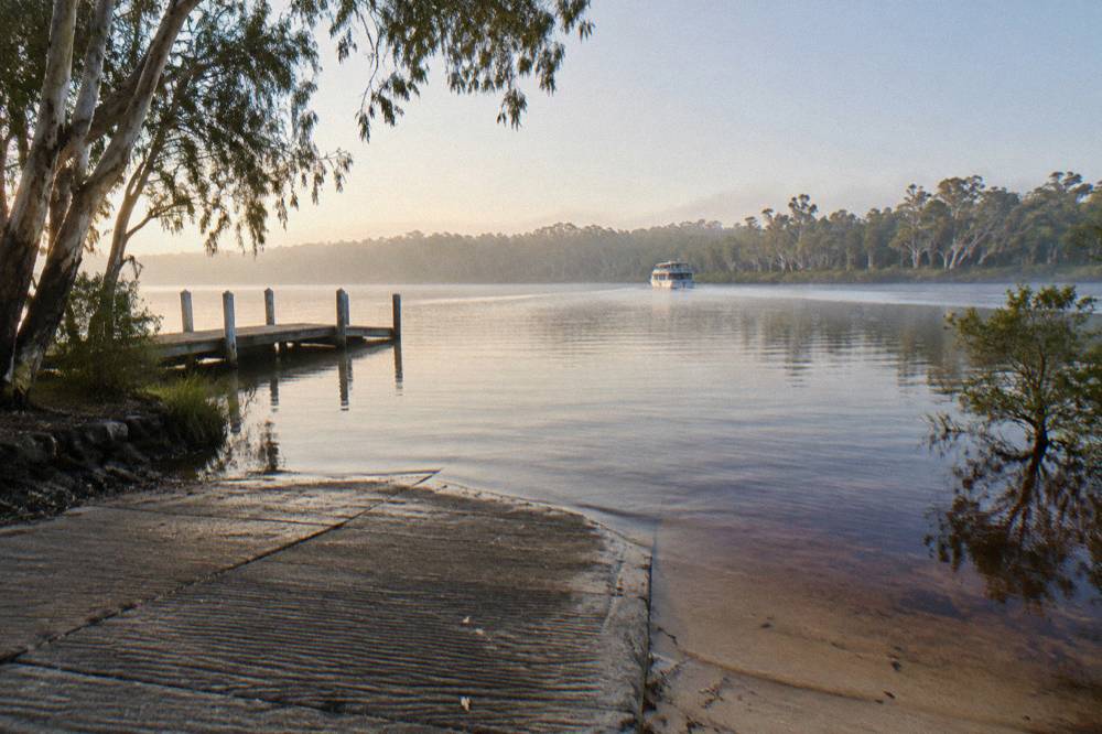 A boat ramp and jetty on tea coloured lake water with paperbark trees along the far shore