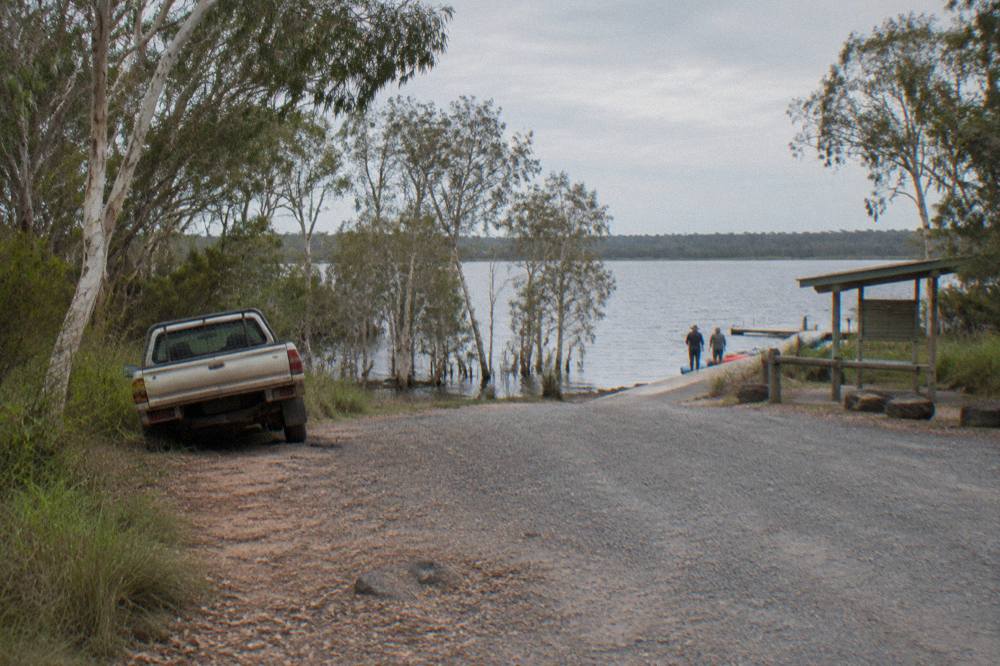 A quiet gravel car park edge beside a lake with paperbark trees at the waterline