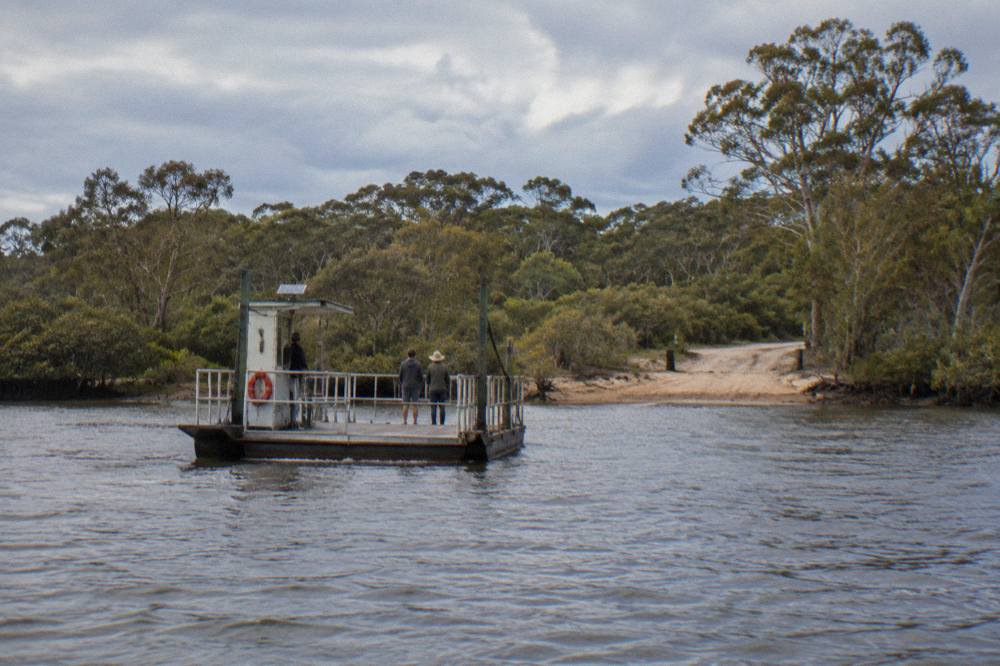 A ferry crossing a wide river channel with a sandy shoreline and distant bushland