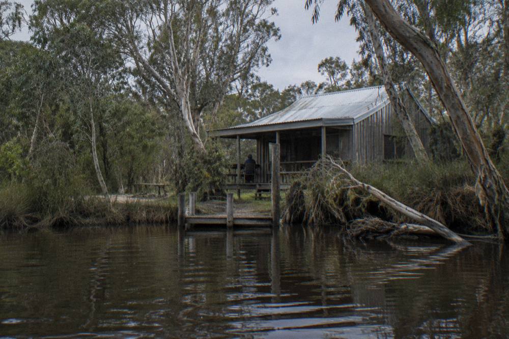 Weathered timber hut beside a quiet tannin river with paperbark trees