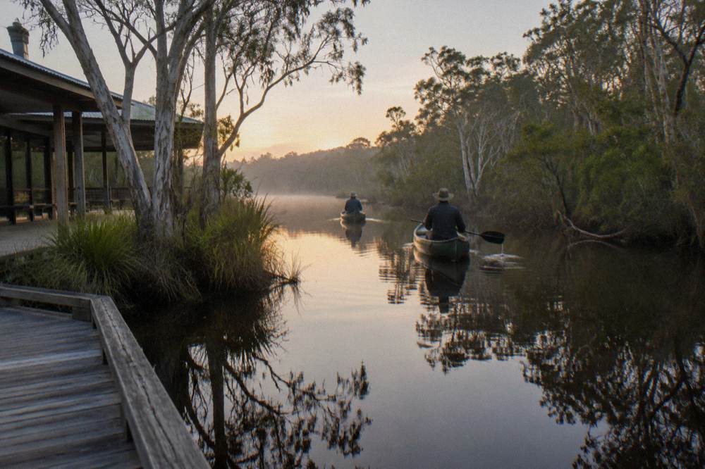 Two canoes floating on mirror-still dark water between forested banks