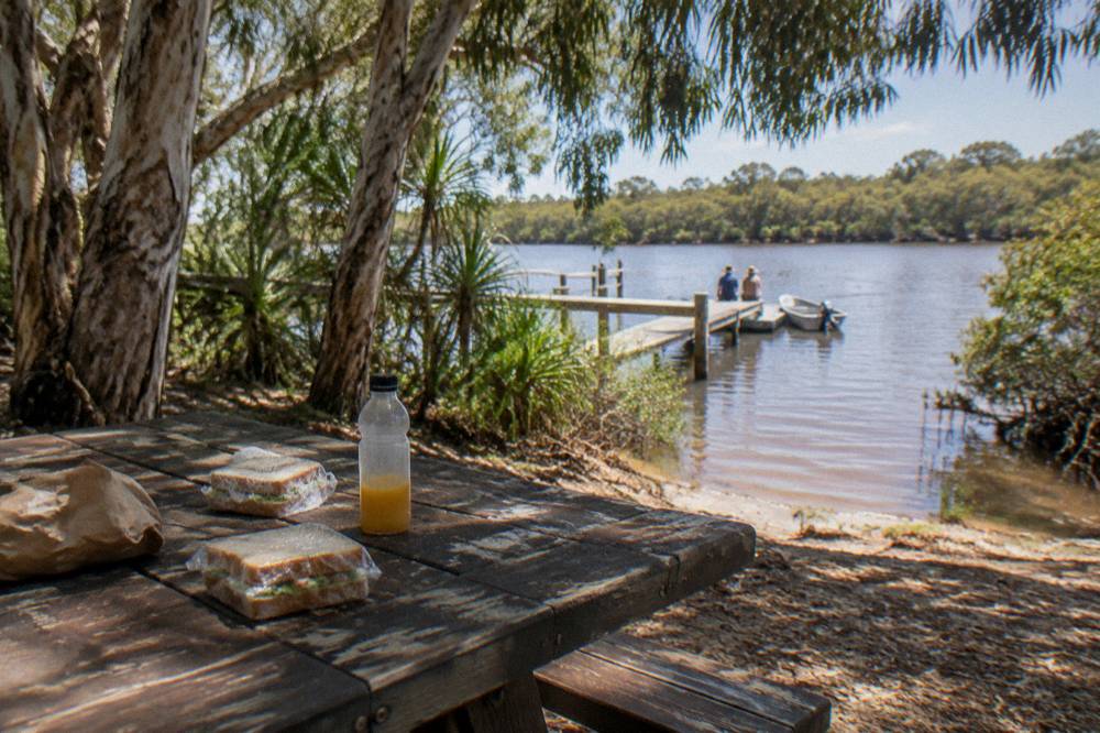 A picnic table set with lunch beside a river jetty under paperbark trees