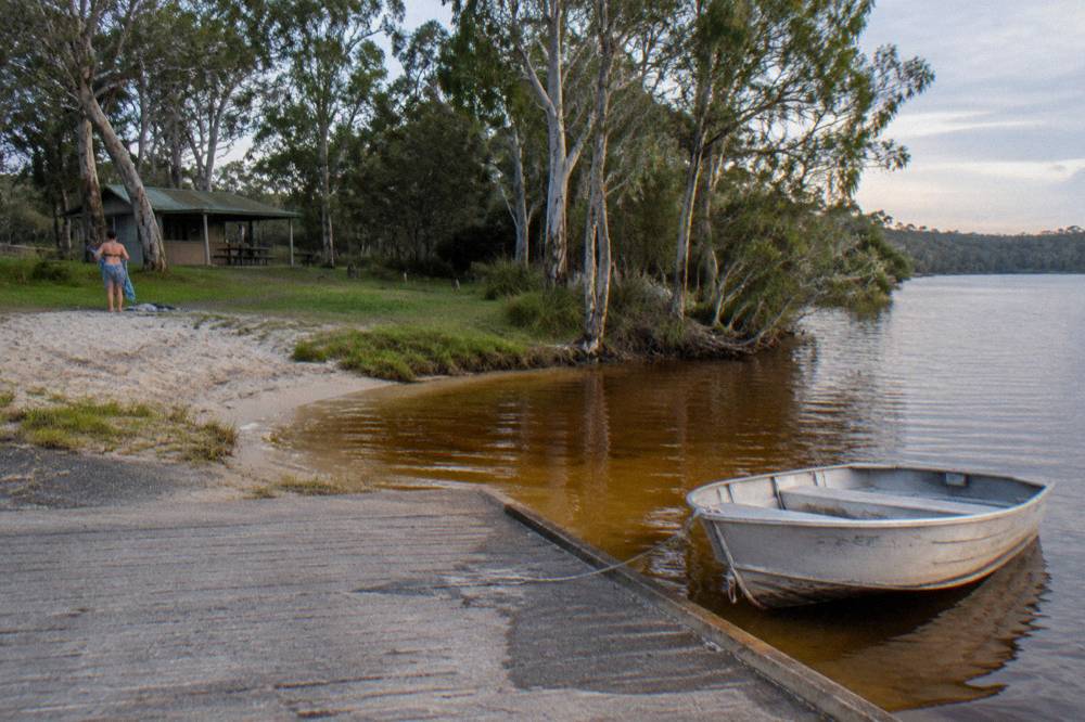 Grassy lake shore with a small boat ramp and still tea coloured water at the edge of a camping area