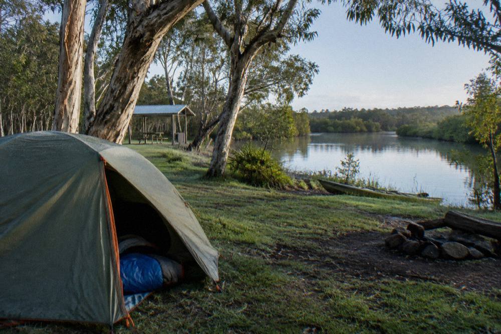 Tent pitched on grass beside a calm river with paperbark trees