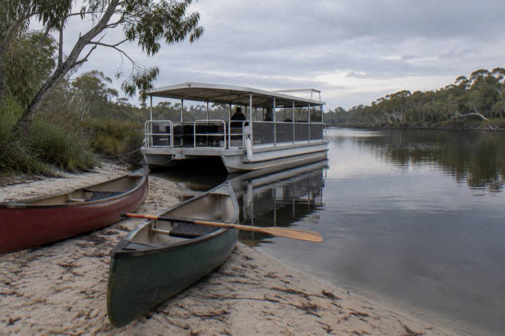 Canoes resting on a riverbank beside a small cruise boat on calm dark water