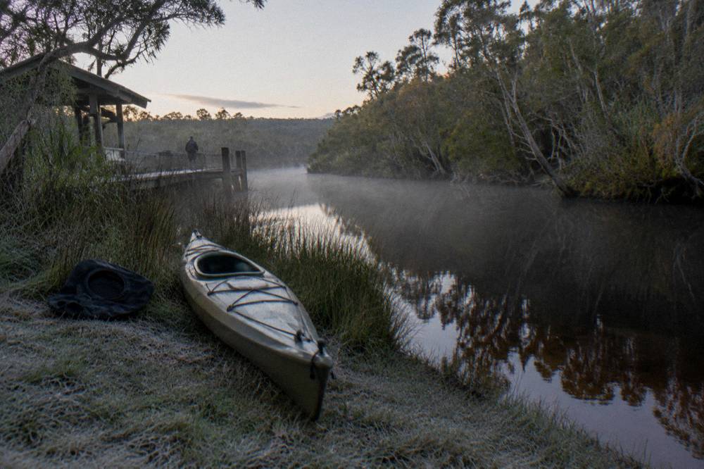A kayak resting on a grassy bank beside dark calm river water in soft morning light